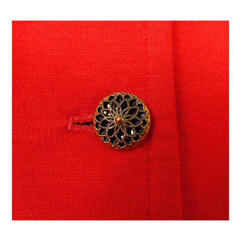 May include: A close-up of a gold-coloured button with a black centre and a decorative design. The button is on a red fabric background.