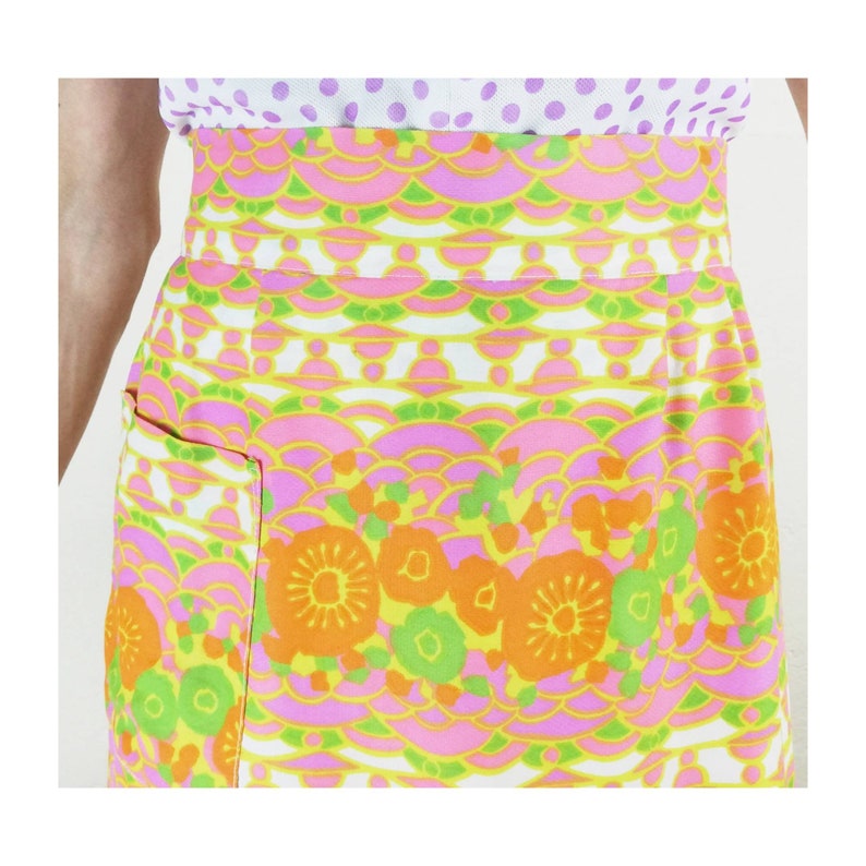 May include: A vintage apron with a colorful floral and geometric pattern in shades of pink, orange, green, and yellow. The apron has a large pocket on the front.