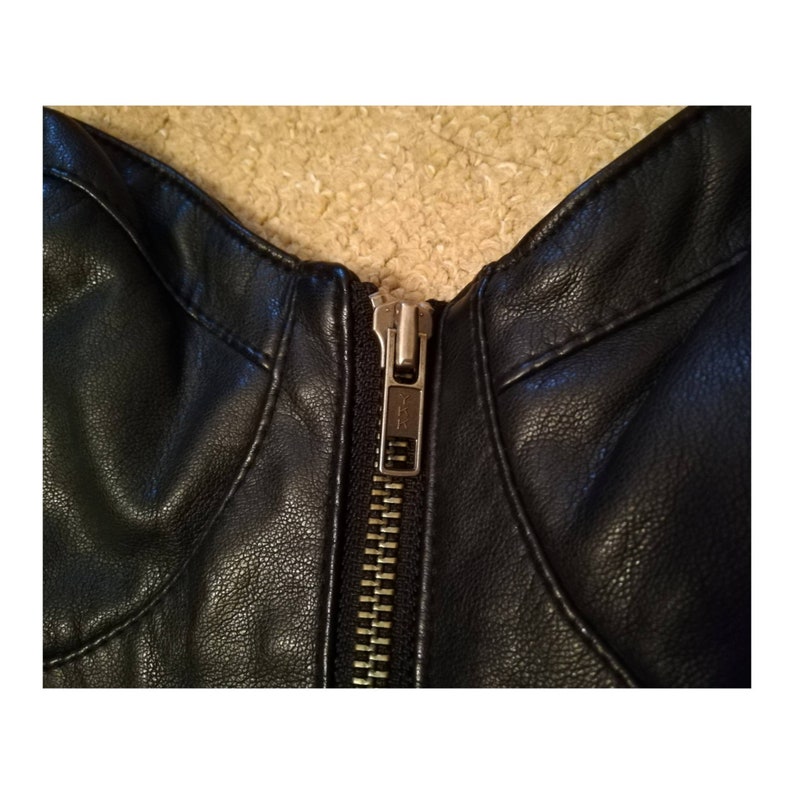 May include: Close-up of a black leather jacket zipper. The zipper is silver with a black pull tab.