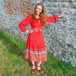 Peut inclure: Une femme portant une robe maxi rouge à imprimé floral et une ceinture marron. Elle se tient devant un mur en pierre.