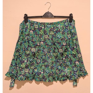 Puede incluir: Falda con estampado floral verde y azul marino colgada de una percha negra. La falda tiene un dobladillo con volantes y detalles de lazos. La tela presenta un patrón repetido de flores y hojas estilizadas en tonos verdes, azules y blancos.