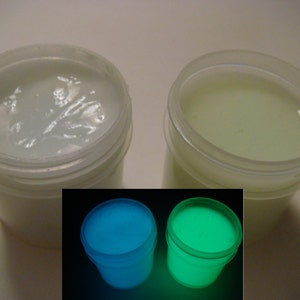 May include: Two white plastic jars of glow-in-the-dark paint. The jars are sitting on a white surface. The paint in the jars is white, but it glows blue and green in the dark.