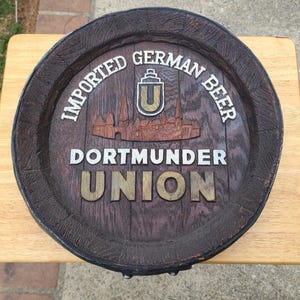 May include: A circular wooden beer barrel head sign featuring the text "IMPORTED GERMAN BEER" and "DORTMUNDER UNION" in raised white and gold lettering. The center displays a carved cityscape graphic and a shield logo with the letter U.
