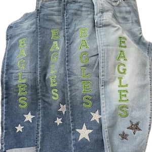 May include: Three pairs of blue denim jeans with the word "EAGLES" spelled vertically in green rhinestones on the side of each leg. Each pair has silver glitter stars on the legs.
