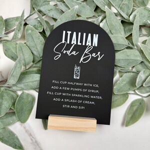 May include: Black and white sign with a rounded top that says "ITALIAN Soda Bar". The sign has instructions for making an Italian soda: "FILL CUP HALFWAY WITH ICE, ADD A FEW PUMPS OF SYRUP, FILL CUP WITH SPARKLING WATER, ADD A SPLASH OF CREAM, STIR AND SIP!"