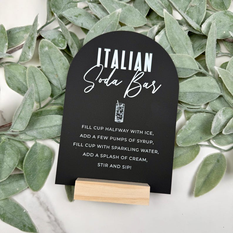 May include: Black and white sign with a rounded top that says "ITALIAN Soda Bar". The sign has instructions for making an Italian soda: "FILL CUP HALFWAY WITH ICE, ADD A FEW PUMPS OF SYRUP, FILL CUP WITH SPARKLING WATER, ADD A SPLASH OF CREAM, STIR AND SIP!"