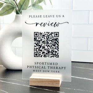 May include: A frosted acrylic sign with a black and white QR code, the text "Please leave us a review" in a cursive font, and the text "Sportsmed Physical Therapy West New York" in a sans-serif font. The sign is on a wooden stand.