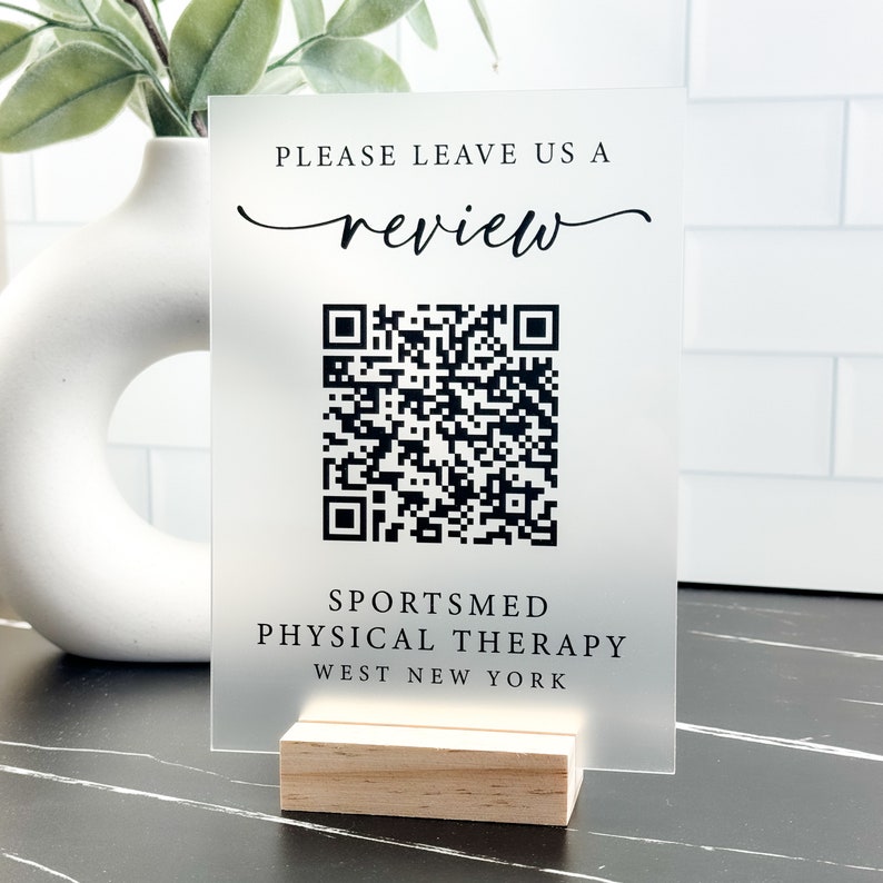 May include: A frosted acrylic sign with a black and white QR code, the text "Please leave us a review" in a cursive font, and the text "Sportsmed Physical Therapy West New York" in a sans-serif font. The sign is on a wooden stand.