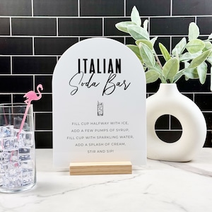 May include: White sign with a black arch at the top that says "ITALIAN Soda Bar".  The sign has instructions for making an Italian soda: "FILL CUP HALFWAY WITH ICE, ADD A FEW PUMPS OF SYRUP, FILL CUP WITH SPARKLING WATER, ADD A SPLASH OF CREAM, STIR AND SIP!"
