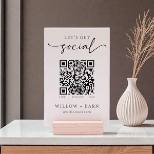 May include: A white sign with the text "LET'S GET social" and a QR code. Below the code are the words "LIKE FOLLOW TAG" and the business name "WILLOW + BARN @willowandbarn". The sign is displayed on a wooden block.