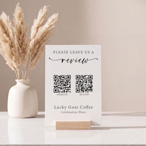 Könnte beinhalten: Ein weißes rechteckiges Schild mit schwarzem Text, der PLEASE LEAVE US A REVIEW, Lucky Goat Coffee und Celebration Place lautet. Es enthält zwei schwarze QR-Codes mit der Beschriftung WEBSITE und REVIEW und steht auf einem Holzsockel.