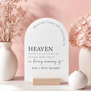 May include: An arched white memorial sign with black text that reads: BECAUSE THOSE WE LOVE ARE IN HEAVEN THERE'S A LITTLE BIT OF HEAVEN HERE TODAY in loving memory of Jack + Betty Spangler. The sign is supported by a rectangular wooden base.
