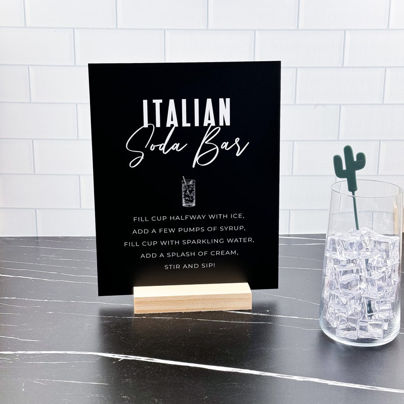 May include: Black sign with white text that reads "ITALIAN Soda Bar" and instructions for making a soda drink. The sign is on a wooden stand. A glass with ice and a green cactus stirrer is in the background.