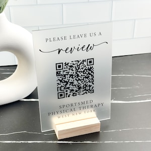 May include: A frosted acrylic sign with a black QR code and the text "Please leave us a review" and "Sportsmed Physical Therapy West New York". The sign is on a wooden stand.
