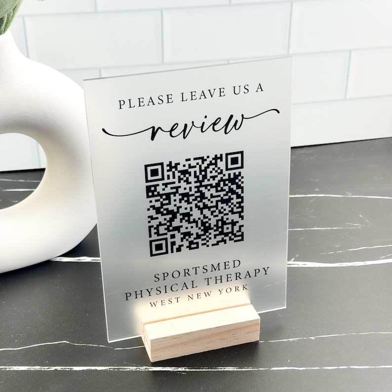 May include: A frosted acrylic sign with a black QR code and the text "Please leave us a review" and "Sportsmed Physical Therapy West New York". The sign is on a wooden stand.