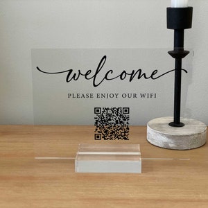 Puede incluir: Un cartel transparente de acrílico con el texto "welcome" en letra cursiva. Debajo del texto está la frase "Please enjoy our wifi" y un código QR en blanco y negro. El cartel está en un soporte de madera.