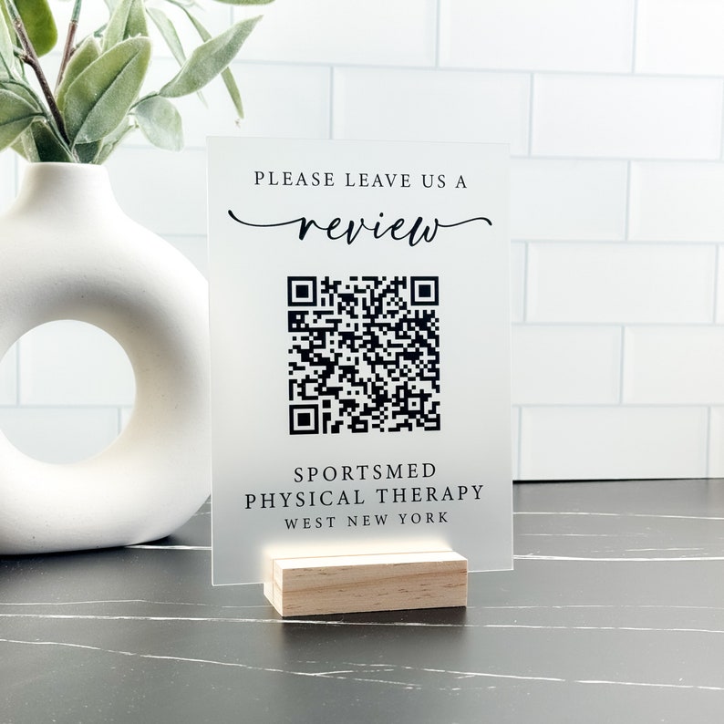 May include: A clear acrylic sign with a black QR code and the text "Please leave us a review" in a cursive font. Below the QR code is the text "SPORTSMED PHYSICAL THERAPY WEST NEW YORK". The sign is on a small wooden stand.