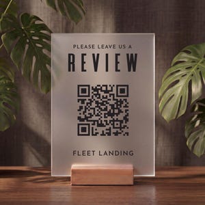 Pode incluir: Uma placa retangular de acrílico fosco sobre uma base de madeira clara. A placa apresenta texto preto que diz PLEASE LEAVE US A REVIEW, um código QR preto central e o texto FLEET LANDING na parte inferior, impressos em uma superfície semitransparente.