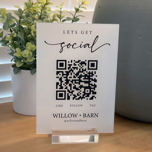 以下が含まれることがあります： 白地に黒文字で「Let's Get Social」と書かれた看板で、その下に白黒のQRコードがあります。「Like Follow Tag」という文字がQRコードの下にあり、「Willow + Barn @willowandbarn」という文字が看板の下部に書かれています。