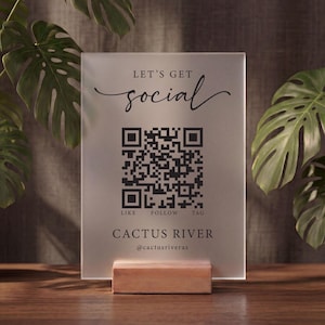 Puede incluir: Un letrero rectangular de acrílico esmerilado con el texto negro "LET'S GET social" en la parte superior, un código QR negro central, las palabras "LIKE FOLLOW TAG" debajo y "CACTUS RIVER" con "@cactusriveraz" al pie, sobre una base de madera.