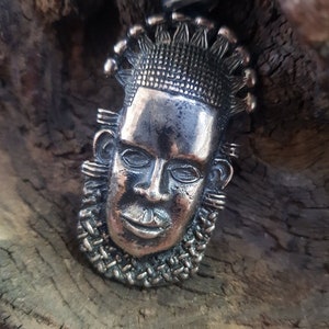May include: A silver pendant featuring a detailed face with a crown of spikes. The pendant is on a wooden surface.