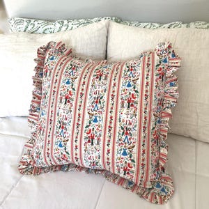 May include: A decorative pillow with a white background and a repeating pattern of red stripes and festive figures. The pillow has a ruffled edge and is placed on a white bed. The pattern includes figures in red, blue, and green.