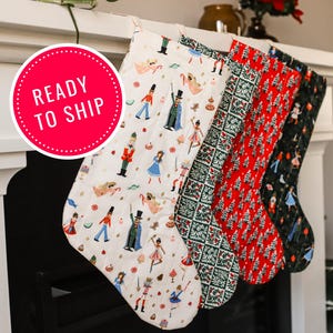 May include: Four Christmas stockings with festive patterns hang from a white mantel. The stockings feature designs with nutcrackers, dancers, and Christmas trees in red, green, and black. A red circle with the text "READY TO SHIP" is visible.