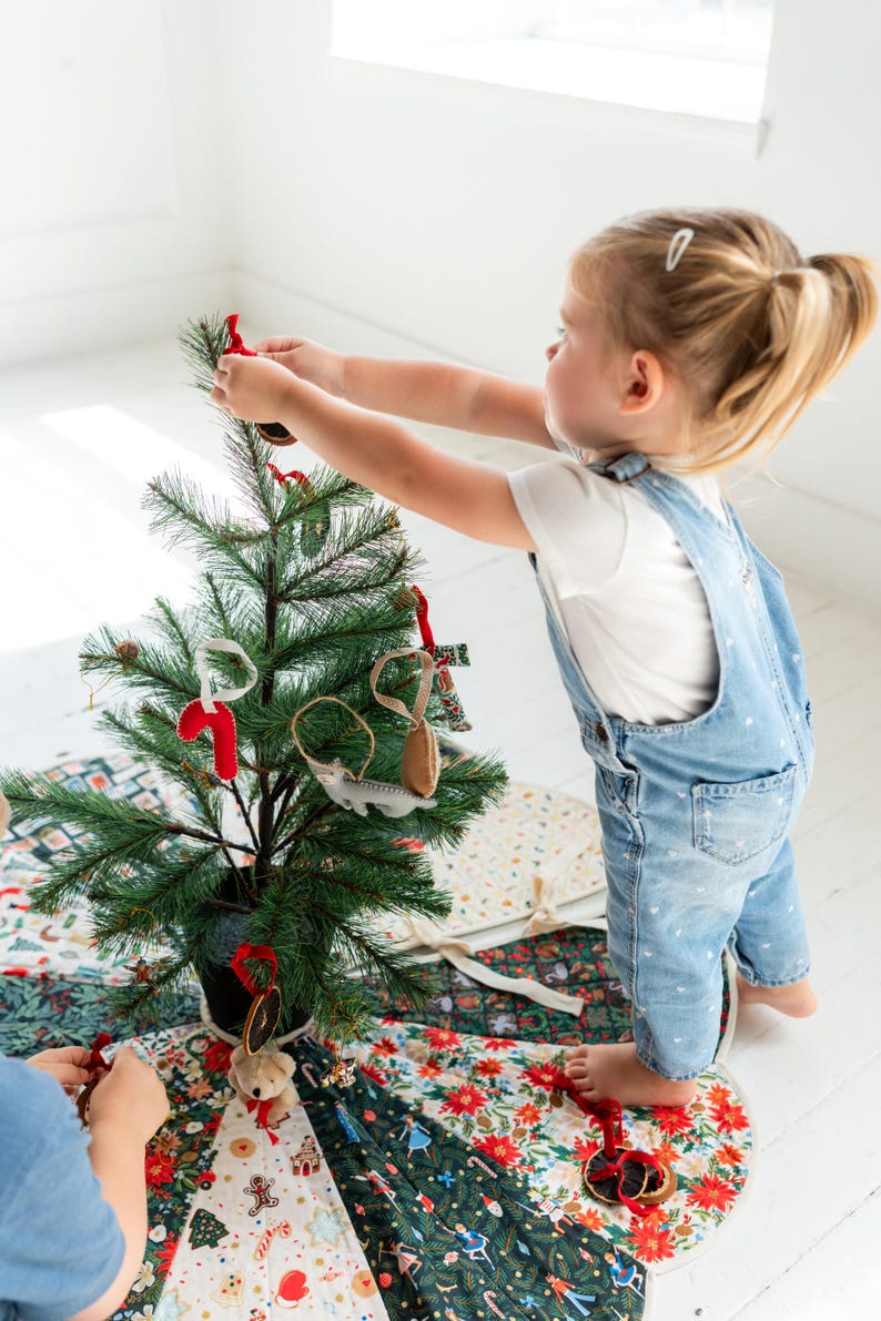 May include: A small artificial green pine tree decorated with felt ornaments including a red candy cane, a gray animal, and dried orange slices. The tree sits on a circular fabric tree skirt featuring a colorful pattern of holiday motifs.