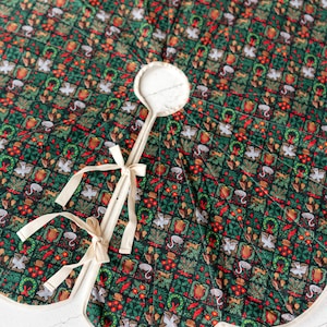 May include: A circular quilted tree skirt featuring a dark green background with a repeating grid pattern of festive motifs including white doves, red tulips, green wreaths, and small animals, finished with cream-colored fabric ties and trim.