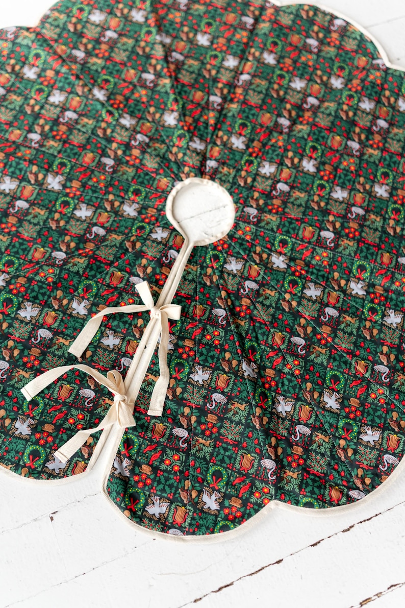 May include: A circular quilted tree skirt featuring a dark green background with a repeating grid pattern of festive motifs including white doves, red tulips, green wreaths, and small animals, finished with cream-colored fabric ties and trim.
