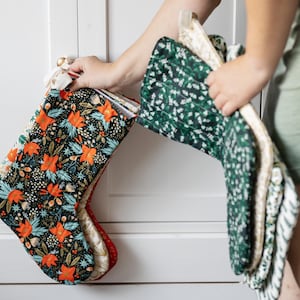 May include: Christmas stockings in various floral patterns. One stocking features a dark background with orange and teal flowers. Another has a green floral design. The stockings are held by a person in a green dress.