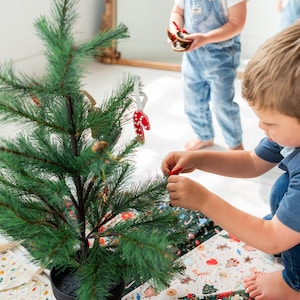 May include: A small artificial green Christmas tree in a black pot, decorated with small ornaments including a red and white candy cane. The tree sits on a circular fabric tree skirt featuring a colorful patchwork pattern of holiday motifs.