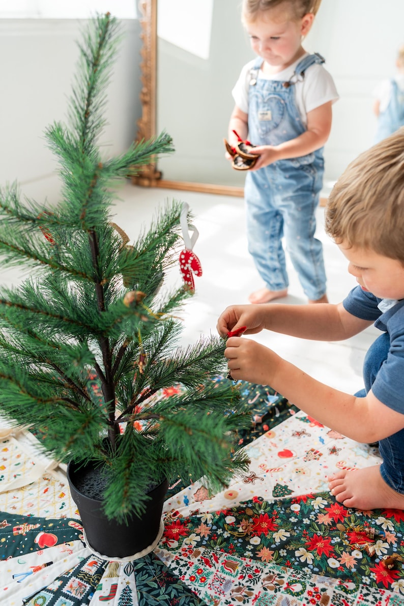 May include: A small artificial green Christmas tree in a black pot, decorated with small ornaments including a red and white candy cane. The tree sits on a circular fabric tree skirt featuring a colorful patchwork pattern of holiday motifs.