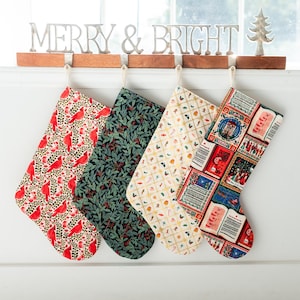 May include: Four fabric Christmas stockings hanging from a wooden rack with a metal MERRY & BRIGHT sign. The stockings feature patterns of red cardinals, dark green foliage, small colorful holiday icons, and vintage-style postage stamps.