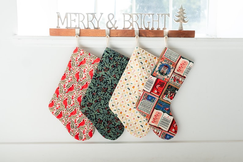 May include: Four fabric Christmas stockings hanging from a wooden rack with a metal MERRY & BRIGHT sign. The stockings feature patterns of red cardinals, dark green foliage, small colorful holiday icons, and vintage-style postage stamps.