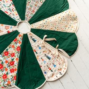 May include: A circular quilted tree skirt with a scalloped edge, featuring alternating panels of solid dark green fabric and cream-colored fabric printed with festive holiday motifs including nutcrackers, poinsettias, and ornaments, secured with cream ties.