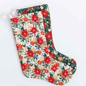May include: Two Christmas stockings featuring a floral pattern with red poinsettias, green pine needles, and small pinecones. One stocking has a white background, while the other has a dark green background. Both include a small fabric hanging loop.