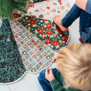 May include: A circular fabric tree skirt featuring alternating panels of dark green foliage patterns, white fabric with small holiday-themed illustrations, and vibrant red poinsettia floral prints, finished with a light-colored piped edge.