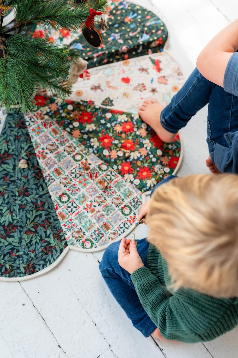 May include: A circular fabric tree skirt featuring alternating panels of dark green foliage patterns, white fabric with small holiday-themed illustrations, and vibrant red poinsettia floral prints, finished with a light-colored piped edge.