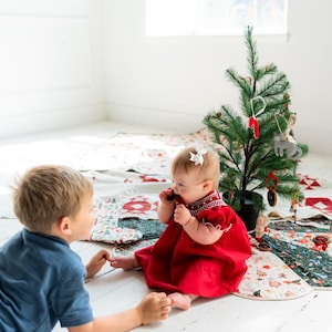 May include: A red dress with white smocked detailing at the neckline and puffed sleeves. The dress features a gathered skirt and is worn by a baby who has a white bow in their hair. The garment is made of a solid red fabric with white stitching.