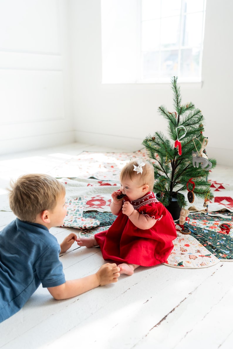 May include: A red dress with white smocked detailing at the neckline and puffed sleeves. The dress features a gathered skirt and is worn by a baby who has a white bow in their hair. The garment is made of a solid red fabric with white stitching.