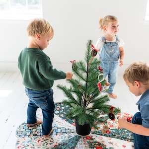 May include: A circular fabric Christmas tree skirt featuring a patchwork design of various holiday-themed patterns in red, green, and white, placed under a small artificial pine tree decorated with ornaments and red ribbon bows.