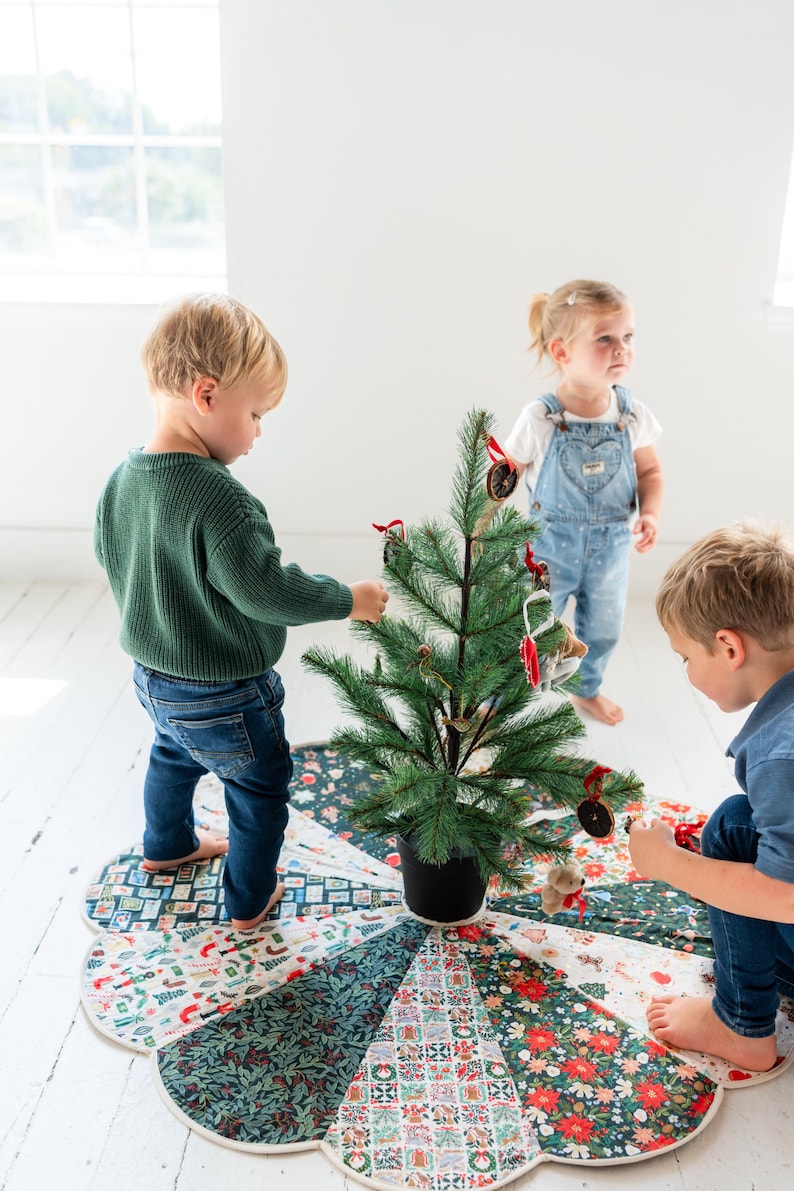 May include: A circular fabric Christmas tree skirt featuring a patchwork design of various holiday-themed patterns in red, green, and white, placed under a small artificial pine tree decorated with ornaments and red ribbon bows.