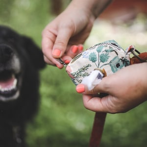 May include: A person holding a small, floral patterned bag with a zipper and a metal clasp. The bag is filled with white wipes. A black dog is in the background.