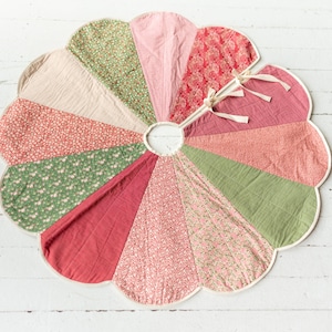 May include: A circular tree skirt featuring a scalloped edge and a central opening with tie closures. The design consists of twelve wedge-shaped panels in alternating shades of sage green, dusty pink, cream, and red, with various floral patterns.