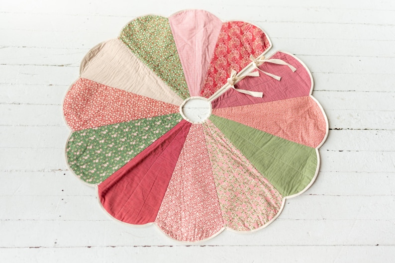May include: A circular tree skirt featuring a scalloped edge and a central opening with tie closures. The design consists of twelve wedge-shaped panels in alternating shades of sage green, dusty pink, cream, and red, with various floral patterns.