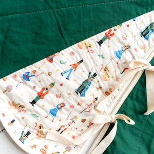 May include: A quilted tree skirt featuring a solid dark green fabric panel and a cream-colored panel with a repeating pattern of nutcrackers, ballerinas, and holiday treats. The skirt includes cream-colored fabric ties for securing the edges.