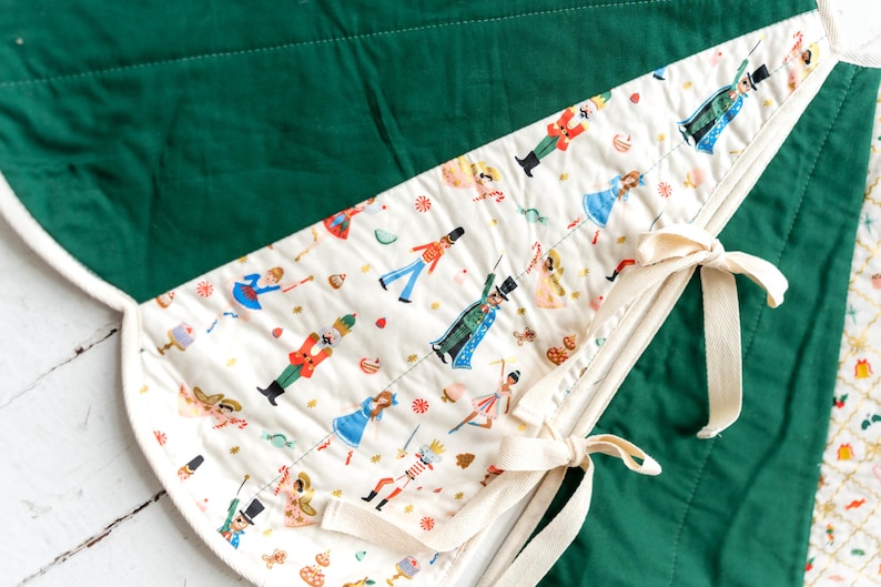 May include: A quilted tree skirt featuring a solid dark green fabric panel and a cream-colored panel with a repeating pattern of nutcrackers, ballerinas, and holiday treats. The skirt includes cream-colored fabric ties for securing the edges.