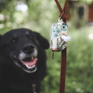 May include: A black dog with a white patch on its chest sits in front of a floral patterned dog waste bag dispenser hanging from a rusty metal hook.