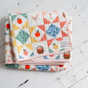 May include: A folded patchwork quilt featuring a repeating pattern of apples, baskets, and chickens on a white background with yellow, blue, and red gingham accents. A white fabric label with the black text TAB Stitchery is sewn on the corner.
